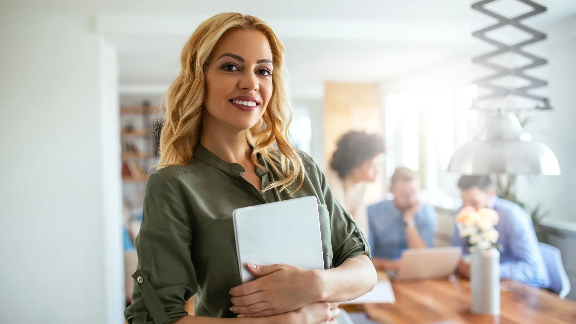 Woman holding her laptop and smiling at the camera with office in background