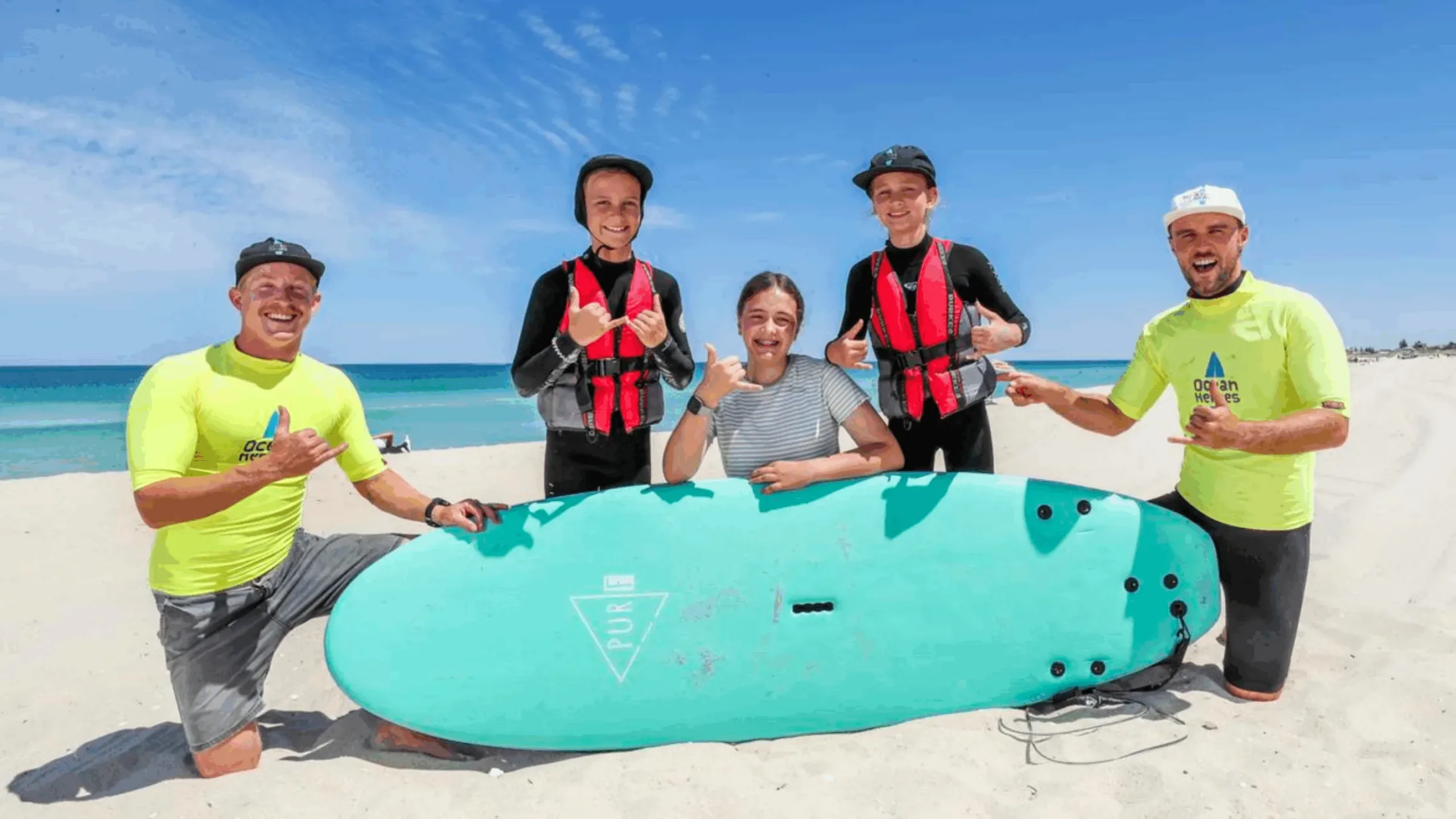 Two surf program volunteers in yellow rash vests with three young participants and a surfboard on a beach