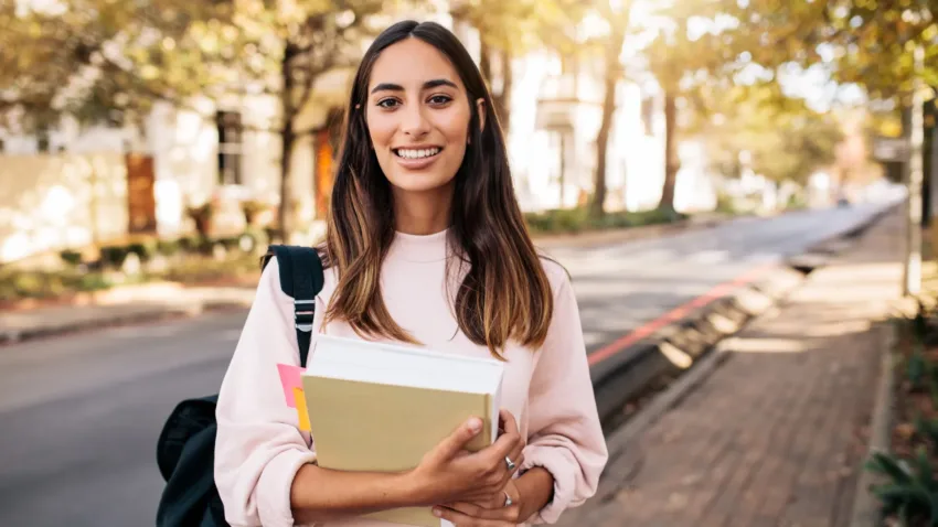 Student holding her notebook and looking at camera with university in background