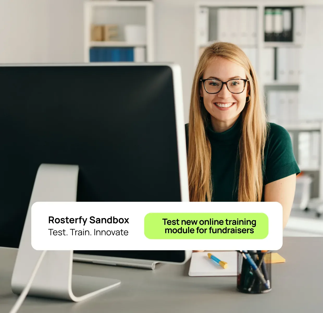 Image of lady smiling at camera working behind her computer - text overlayed to show the Rosterfy Sandbox and the testing of new training for fundraisers