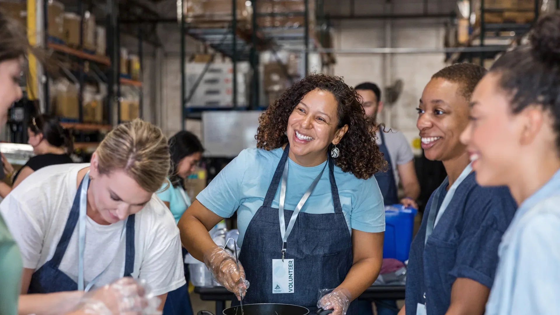 Smiling lady with an apron and lanyard on stirring a big pot and cooking in a kitchen with other volunteers